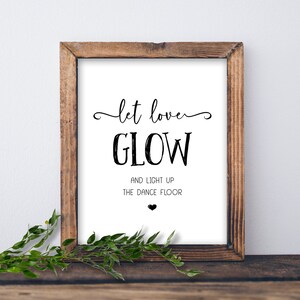May include: A white sign with black text in a wooden frame. The sign says "Let love glow and light up the dance floor" with a small heart at the bottom.