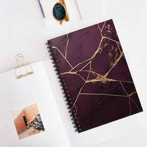 May include: A burgundy spiral notebook with a gold foil design. The design features a cracked marble pattern with gold veins.