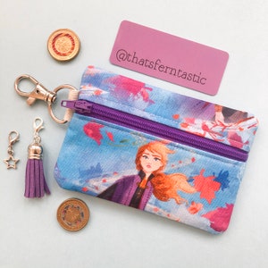 Puede incluir: Un pequeño monedero con cremallera morada y un diseño floral azul y rosa. El monedero presenta un personaje de una popular película de animación. Una borla morada y un dije de estrella cuelgan de un cierre plateado. El texto "@thatsferntastic" está en una etiqueta morada.
