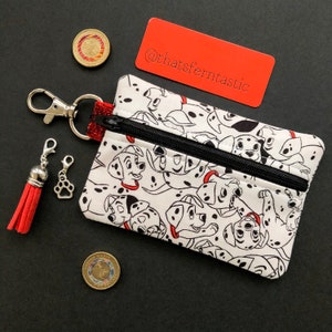 May include: A white zippered pouch featuring a Dalmatian print with black spots and red collars. The pouch has a black zipper, a red tassel, and a silver clasp. A small paw charm and two coins are also visible.