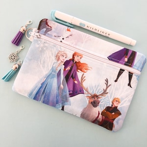 Puede incluir: Dos estuches con cremallera con un tema invernal con Elsa, Anna, Kristoff, Sven y Olaf. Un bolígrafo Mildliner azul claro descansa encima. Borlas y un dije de copo de nieve cuelgan de la cremallera.