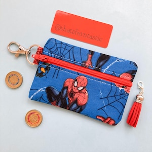 Puede incluir: Un monedero azul con estampado de Spider-Man, cremallera roja y borla. Tiene un mosquetón plateado y una etiqueta roja con el texto "@thatsferntastic".