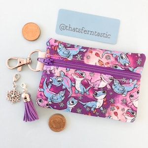 May include: A small, zippered coin purse with a purple and pink pattern featuring blue and white cartoon lizards. The purse has a purple zipper, a silver clasp, and a purple tassel charm. A snowflake charm and two pennies are also visible.