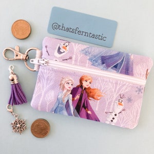Puede incluir: Una pequeña bolsa con cremallera de color lavanda con personajes de la película Frozen, incluyendo a Elsa, Anna y Olaf. La bolsa tiene un cierre plateado, una borla morada y un dije de copo de nieve. El texto "@thatsferntastic" está en una tarjeta azul.