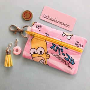 May include: A pink zippered pouch featuring Homer Simpson with Japanese text and a yellow zipper. Includes a gold tassel charm, a pink donut charm, a silver keychain, and a pink tag with the text "@thatsferntastic".