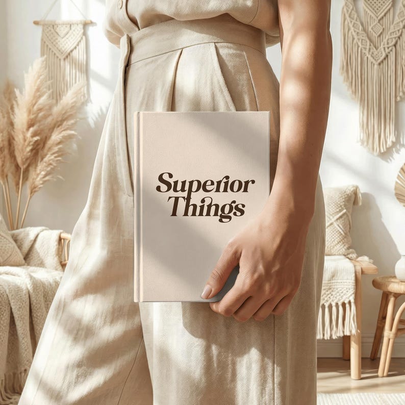 May include: A beige book titled "Superior Things" is held in front of a person. The book's cover is a light beige color with the title in a dark brown, stylized font. The person is wearing beige clothing.