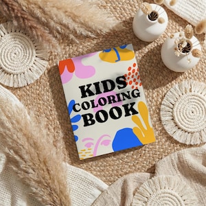 May include: A colorful kids' coloring book with the text "KIDS COLORING BOOK" on the cover. The cover features abstract shapes in pink, blue, yellow, and orange. The book is on a woven surface with decorative elements.