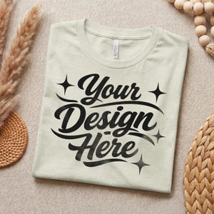 May include: A light green t-shirt with the text "Your Design Here" in black script. The shirt is folded and lies on a neutral-toned surface, with decorative elements in the background.