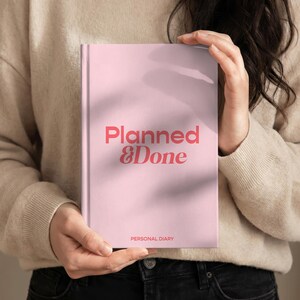 May include: A pink personal diary with the words "Planned & Done" in coral lettering. The diary also has the words "Personal Diary" printed at the bottom. The diary is held by a person wearing a beige sweater and black jeans.