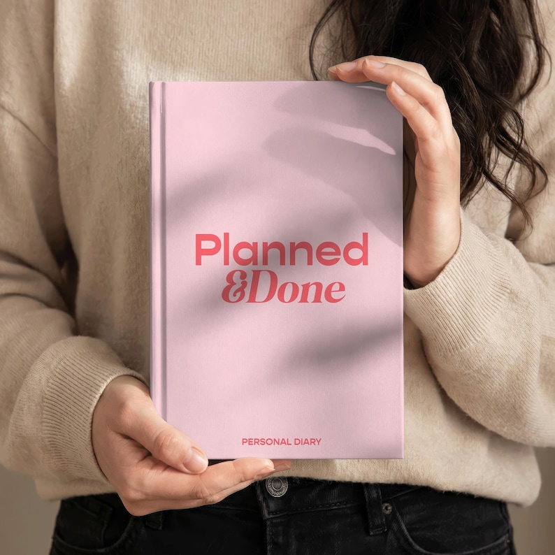 May include: A pink personal diary with the words "Planned & Done" in coral lettering. The diary also has the words "Personal Diary" printed at the bottom. The diary is held by a person wearing a beige sweater and black jeans.