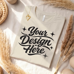 Può includere: Una maglietta color crema con la scritta "Your Design Here" in un carattere nero stilizzato. Un ciondolo a forma di cuore dorato pende dallo scollo. La maglietta è circondata da elementi decorativi, tra cui piante secche e ceramiche.