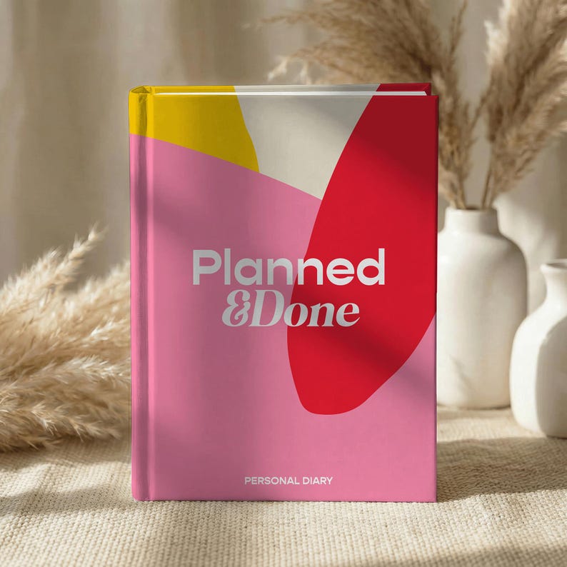 May include: A pink personal diary with the text "Planned & Done" in silver lettering. The cover features a red and yellow abstract design. The diary is standing upright on a textured surface.