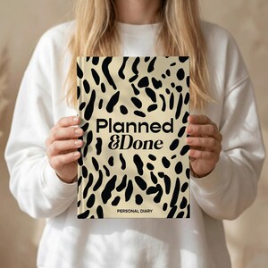 May include: A hardcover personal diary with a cream-colored background and black abstract spots. The words "Planned & Done" are printed in black, with "Personal Diary" below. The diary is held by a person wearing a white sweatshirt.