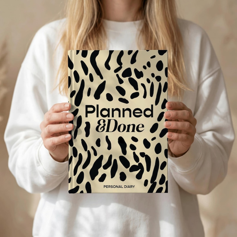 May include: A hardcover personal diary with a cream-colored background and black abstract spots. The words "Planned & Done" are printed in black, with "Personal Diary" below. The diary is held by a person wearing a white sweatshirt.