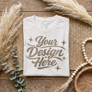 Op de afbeelding: Een wit t-shirt met de tekst "Your Design Here" in een bruin, gestileerd lettertype. Het shirt ligt plat op een geweven mat, met gedroogde planten en decoratieve kralen op de achtergrond. Het shirt is een leeg canvas voor aangepaste ontwerpen.