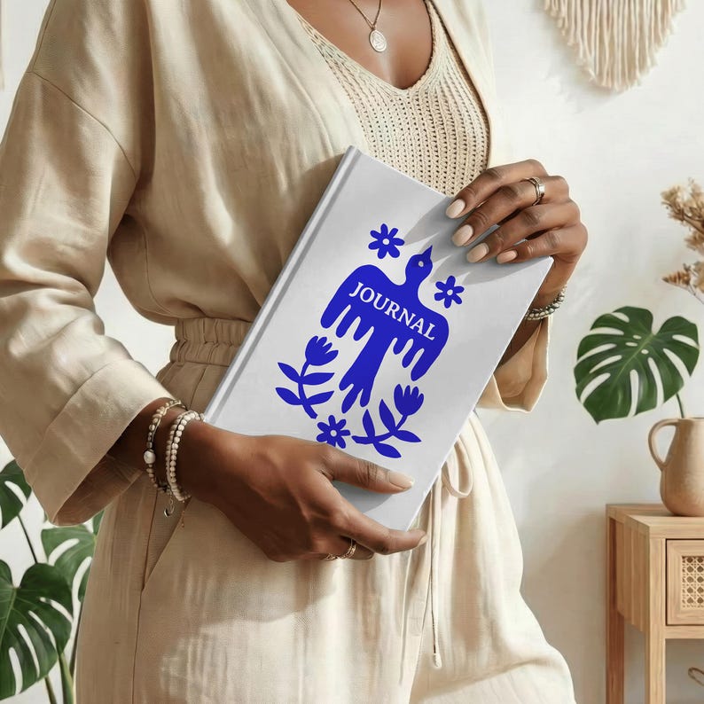 May include: A white journal with a blue bird and floral design. The word "JOURNAL" is printed in blue. The journal is held in front of a person wearing a beige outfit.