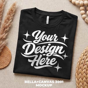 May include: Black t-shirt with the text "Your Design Here" in white script and star accents. The shirt is a Bella+Canvas 3001 mockup, perfect for showcasing custom designs. The shirt is folded neatly on a neutral surface.