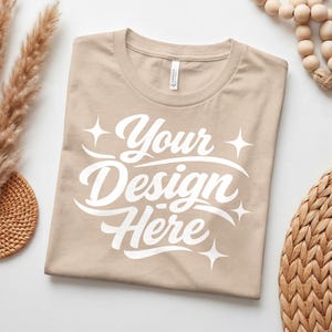 Peut inclure: Un t-shirt beige plié avec le texte "Your Design Here" en écriture blanche. Le t-shirt a un col rond et est fait d'une matière douce. Le design est centré sur le devant du t-shirt. Le t-shirt est une toile vierge pour des designs personnalisés.