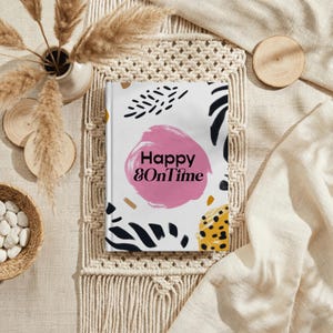 May include: A hardcover journal with the text "Happy & OnTime" on the cover. The cover design features a pink circle, black brushstrokes, and yellow and brown accents. The journal is set on a macrame placemat.