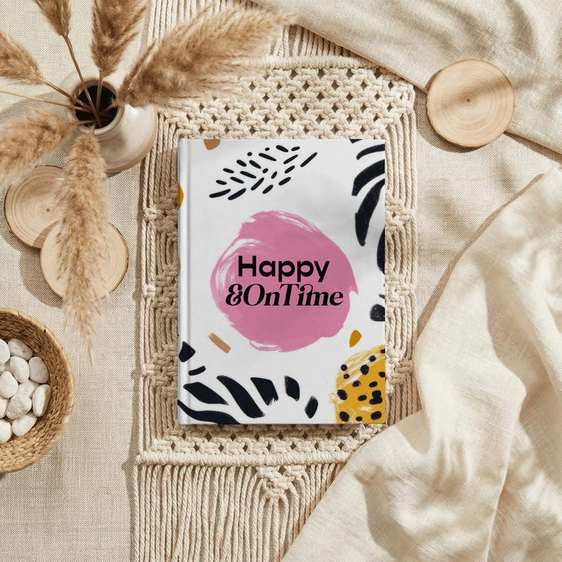 May include: A hardcover journal with the text "Happy & OnTime" on the cover. The cover design features a pink circle, black brushstrokes, and yellow and brown accents. The journal is set on a macrame placemat.
