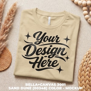 Könnte beinhalten: Ein gefaltetes sandfarbenes T-Shirt mit dem Text "Your Design Here" in schwarzer Schrift. Das Shirt ist ein Bella+Canvas 3001 Mockup. Der Text ist von Sternen umgeben. Das Shirt befindet sich auf einem neutralen Hintergrund.