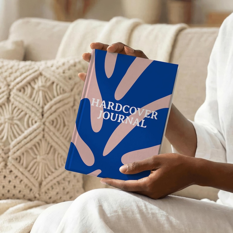 May include: A hardcover journal with a blue cover featuring a pink leaf design. The words "HARDCOVER JOURNAL" are printed in white. The journal is held in someone's hands.