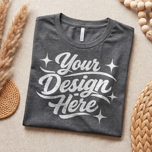 Puede incluir: Una camiseta gris oscuro doblada con el texto "Your Design Here" en escritura blanca. La camiseta está sobre una superficie de tonos neutros con elementos decorativos en el fondo.