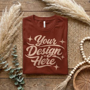 May include: A terracotta-coloured t-shirt featuring the text "Your Design Here" in a cream script. The shirt is displayed on a woven mat, with decorative elements including dried plants and a beaded garland.