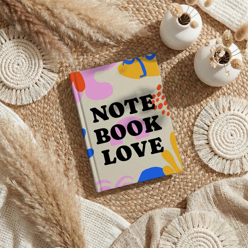 May include: A hardcover notebook with the words "NOTE BOOK LOVE" in black letters. The cover has a beige background with colorful abstract shapes in orange, pink, yellow, and blue. The notebook is on a woven mat with decorative elements.
