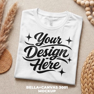 Könnte beinhalten: Ein weißes T-Shirt mit Rundhalsausschnitt und dem Text "Your Design Here" in schwarzer, stilisierter Schrift. Das Bella+Canvas 3001 Mockup wird gefaltet auf einer neutralen Oberfläche präsentiert. Ideal für die Präsentation individueller Designs.