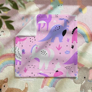 May include: Pink baby blanket with a repeating pattern of purple and white dinosaurs, black dots, and pink accents. The blanket has a white border and is made of a soft material. The blanket is on a pink rug with a rainbow.