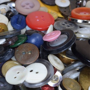 May include: A collection of various buttons in different colors, shapes, and sizes. The buttons are made of different materials, including plastic, metal, and fabric. Some of the buttons have two holes, while others have four holes. The buttons are arranged in a random pattern.