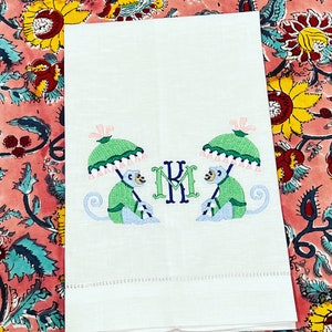 May include: White linen towel with embroidered design. Features two monkeys holding green umbrellas with pink tassels. The initials "KM" are embroidered in green and navy blue. The towel is folded and has a stitched border.