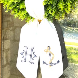 May include: White fabric bow with a blue embroidered letter H and a blue anchor with a gold rope. The bow is tied around a green wreath.