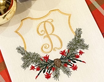 May include: A white linen tea towel with a gold embroidered monogram "B" in a shield shape. The shield is surrounded by a green pine branch with red berries and a brown pine cone.
