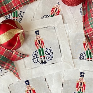 May include: Set of white linen napkins with embroidered nutcracker designs and monogrammed initials. The napkins are decorated with red, green, and blue embroidery.