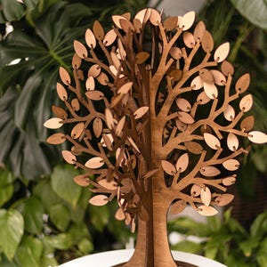 Könnte beinhalten: Eine Holzbaumskulptur mit rundem Sockel und aufwendigen Blattausschnitten. Der Baum besteht aus hellem und dunklem braunem Holz mit natürlichem Finish. Die Skulptur wird auf einer weißen Oberfläche präsentiert, umgeben von grünem Laub.