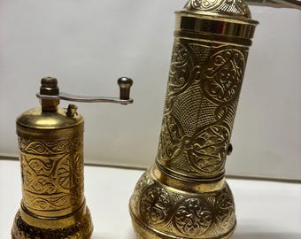 May include: Two antique brass pepper grinders with ornate floral patterns. The grinders have a cylindrical shape with a crank handle on the side. The brass has a warm, golden hue, and the design is etched into the metal.