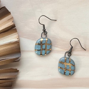 May include: A pair of blue and gold geometric ceramic earrings with a grid pattern. The earrings have a simple hook closure.