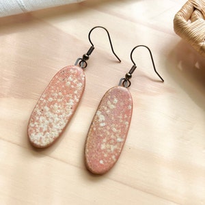 May include: A pair of oval-shaped earrings with a speckled, light pink and white glaze. The earrings have a simple, minimalist design and are made with a bronze-colored metal.