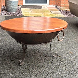 May include: A round wooden top fire pit table with a black metal base and three legs. The table is made of metal and wood and has a rustic look.