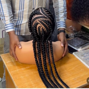 Puede incluir: Una peluca negra con trenzas largas y gruesas, peinadas con un patrón complejo. Las trenzas están dispuestas en un patrón circular en la parte superior de la peluca y luego se extienden hacia abajo en la parte posterior en largos mechones rectos.