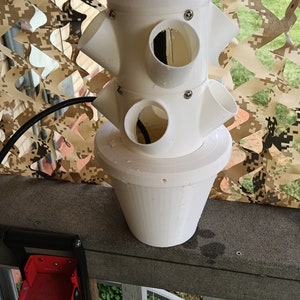 May include: White plastic vertical planter with multiple round openings for planting. The planter is on a wooden railing with a red tool hanging on the railing.