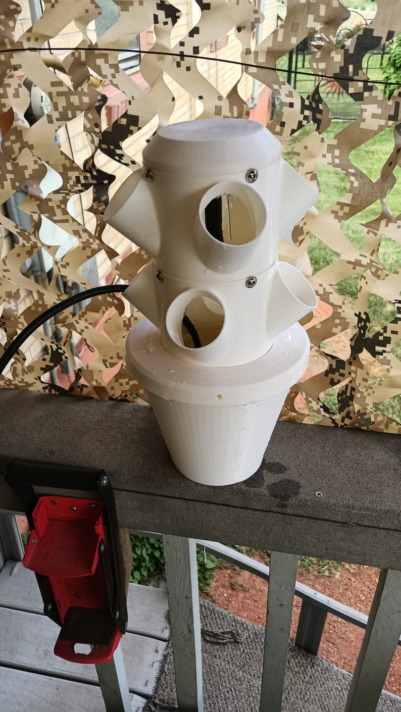 May include: White plastic vertical planter with multiple round openings for planting. The planter is on a wooden railing with a red tool hanging on the railing.