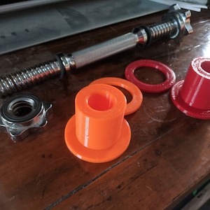 以下が含まれることがあります： ねじ切り端と星形ナットが付いた銀色のダンベルのクローズアップ。ダンベルは木製の表面に置かれ、2組のウェイトプレートがあります。1組はオレンジ色、もう1組は赤色です。