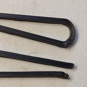May include: Two black plastic clips, each with a single loop, are shown on a white surface. The clips are made of multiple layers of plastic, giving them a textured appearance.
