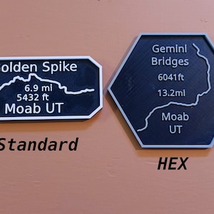 May include: Two black and white signs with text. One is rectangular and reads "Golden Spike" with distance and elevation details. The other is a hexagon and reads "Gemini Bridges" with distance and elevation details. Both signs say "Moab UT".