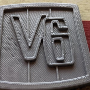 May include: A gray plastic plaque with the letters "V6" embossed on it. The plaque has a textured surface and a raised border.