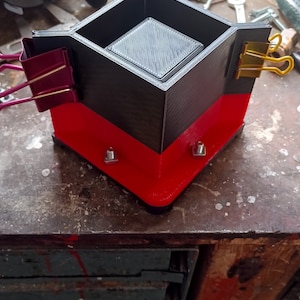 Op de afbeelding: Een 3D-geprinte zwarte en rode doos met een vierkante opening aan de bovenkant. De doos wordt bij elkaar gehouden met twee gouden binderclips en twee rode binderclips. De doos staat op een houten oppervlak.