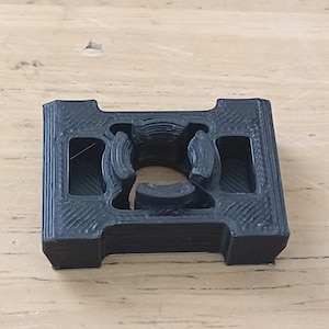 Op de afbeelding: Een zwart, 3D-geprint plastic object met een rechthoekige vorm en een centrale opening. Het object heeft een getextureerd oppervlak en lijkt een onderdeel of deel van een grotere assemblage te zijn. Het ontwerp bevat afgeronde en hoekige kenmerken.